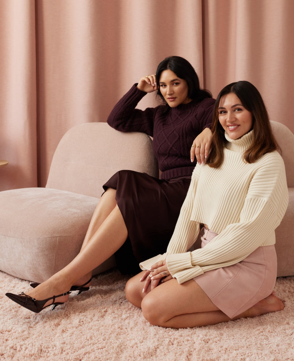 Zwei Frauen in Strickpullovern sitzen auf einem rosa Sofa vor einem rosa Vorhang und lächeln in die Kamera.