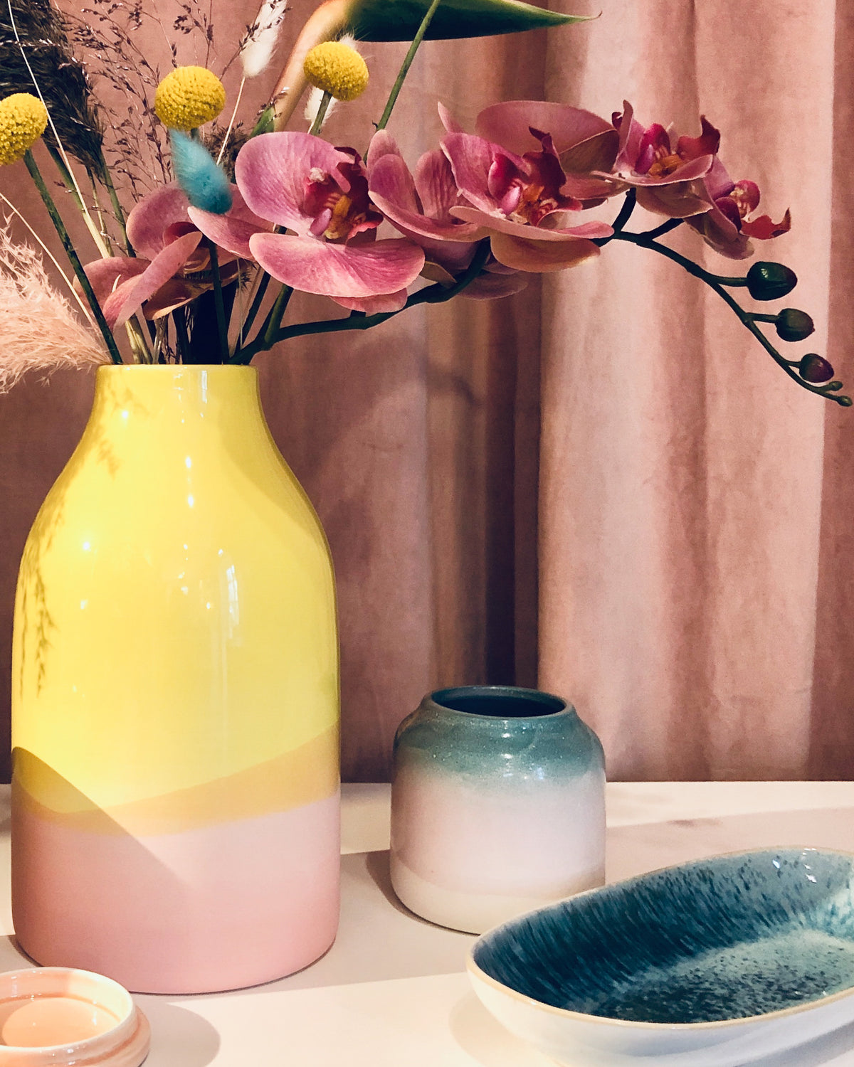 Ein gelb-pinkes Vasenarrangement mit rosa Orchideen vor einem rosafarbenen Vorhang.