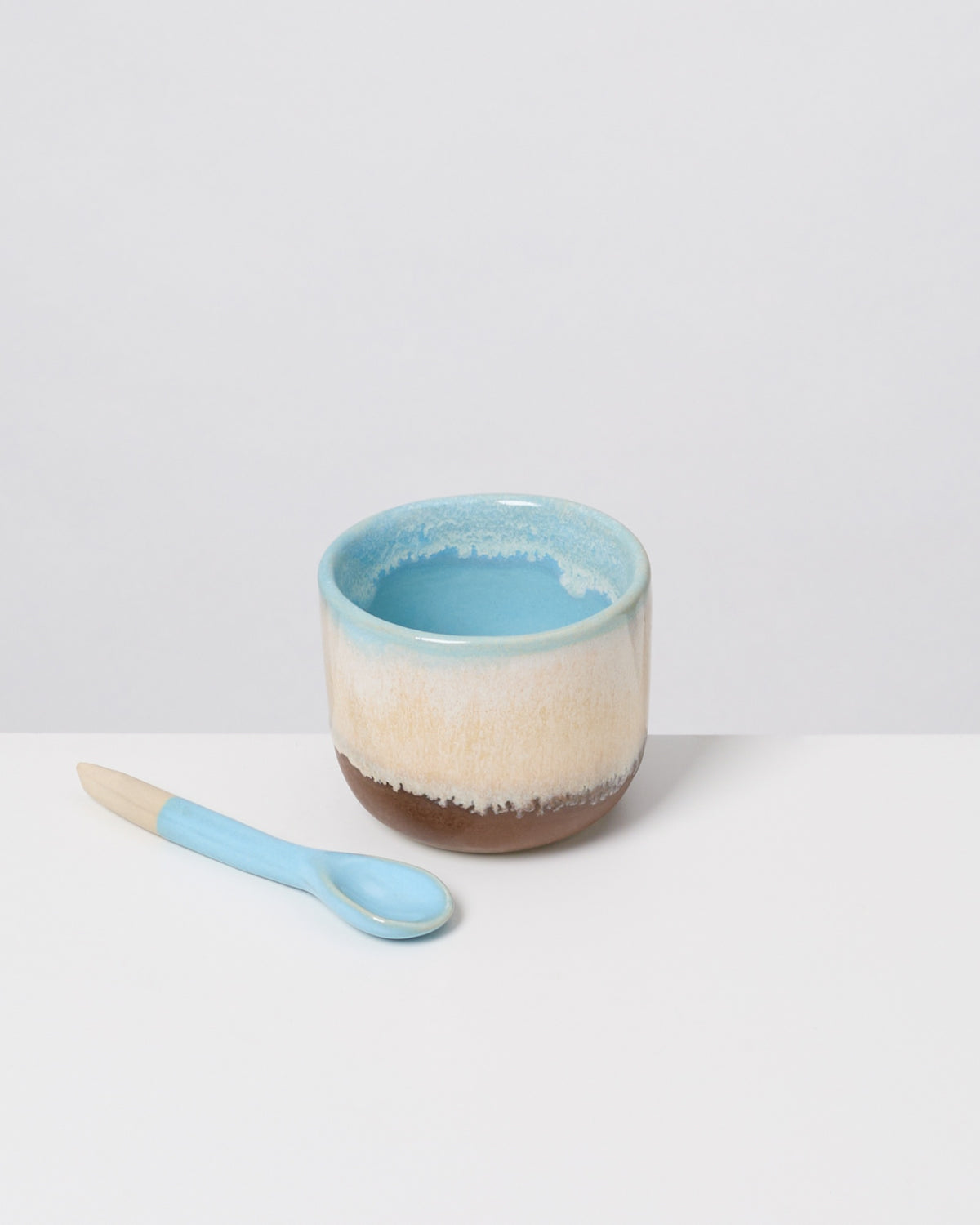 Keramikbecher mit blauem Inneren und passendem Löffel auf weißem Hintergrund.