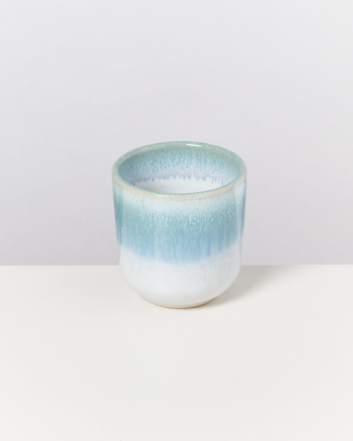 Kleine Keramikvase in Türkis und Weiß mit glatter Oberfläche steht auf einem weißen Tisch vor einem neutralen Hintergrund.