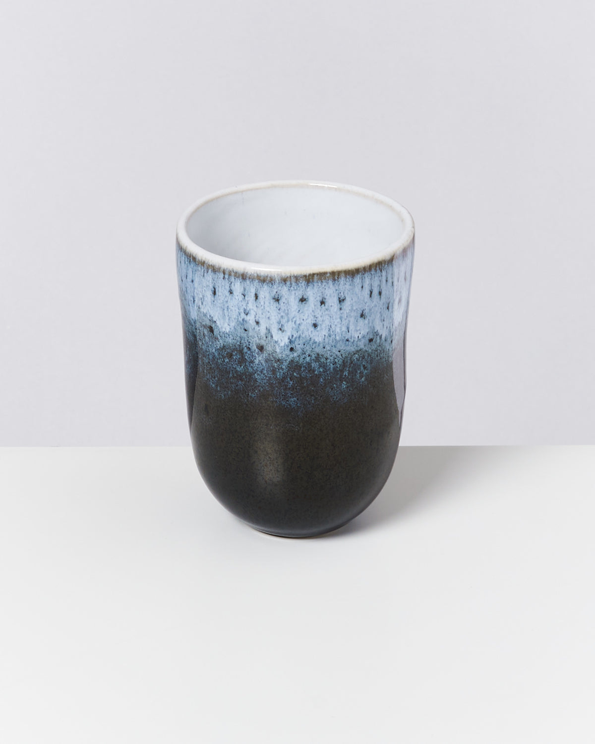 Keramiktasse mit glattem, dunklem Finish und blau-weißer, gesprenkelter Glasur am oberen Rand auf hellem Hintergrund.