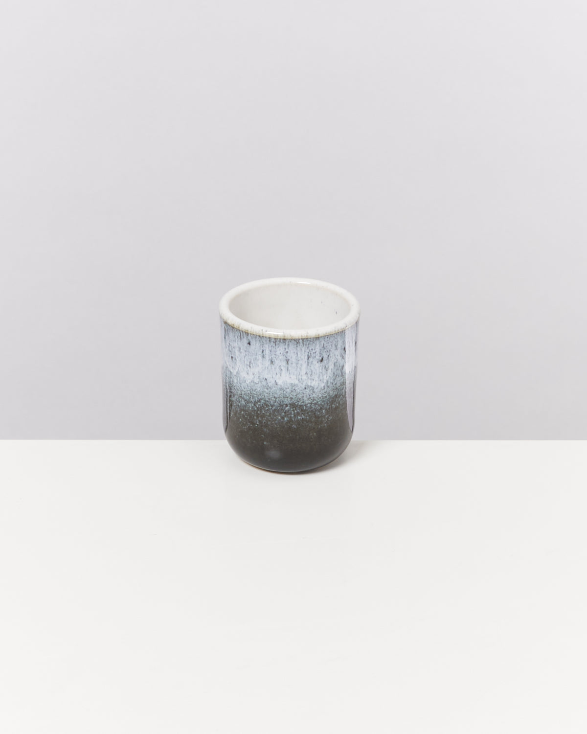 Ein kleiner Becher mit blau-weißer Glasur steht auf einem weißen Tisch vor einem grauen Hintergrund.