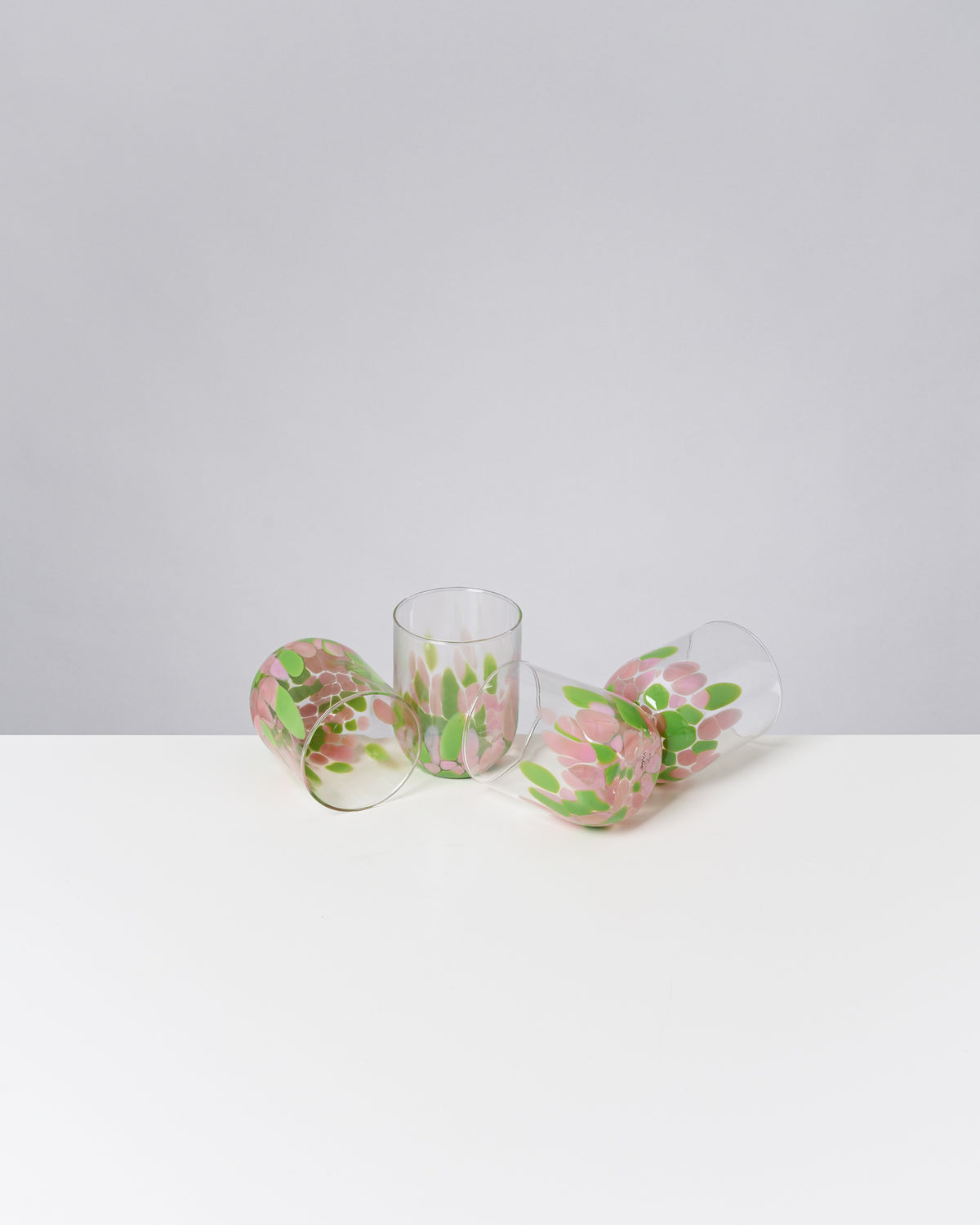 Drei gläserne Becher mit rosa und grünen Mustern liegen auf einem weißen Tisch vor grauem Hintergrund.