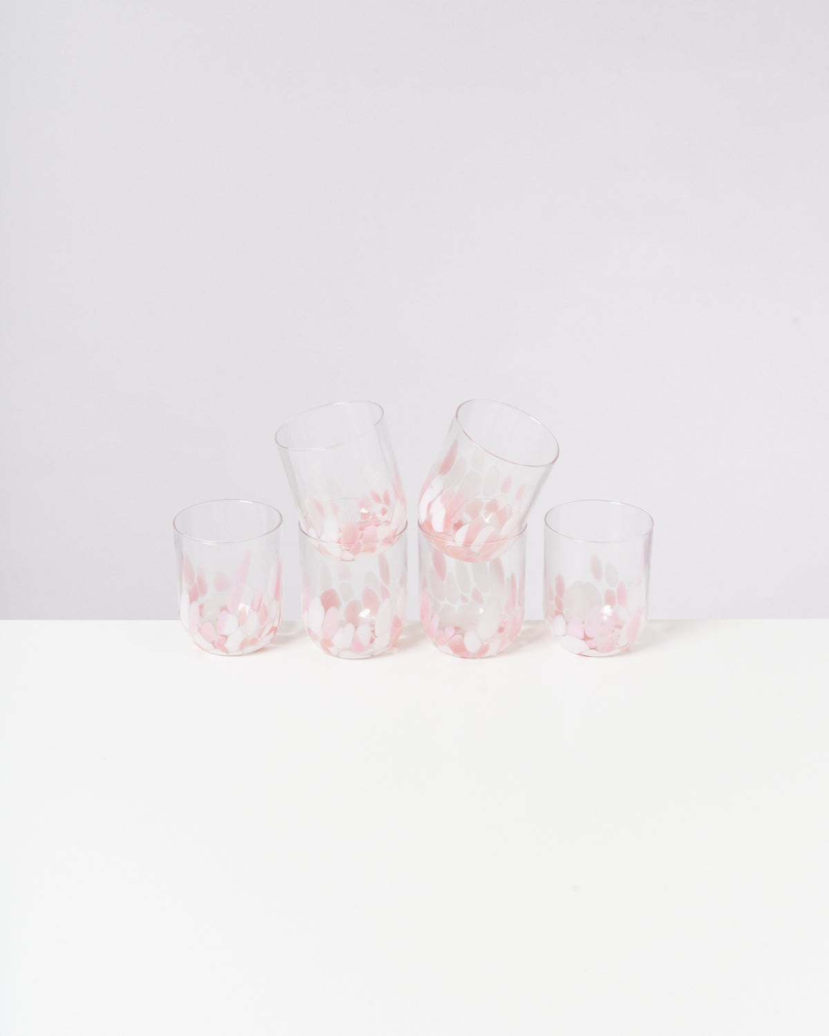 Fünf transparente Gläser mit rosa-weißen Mustern stehen auf einem weißen Tisch vor einer grauen Wand.