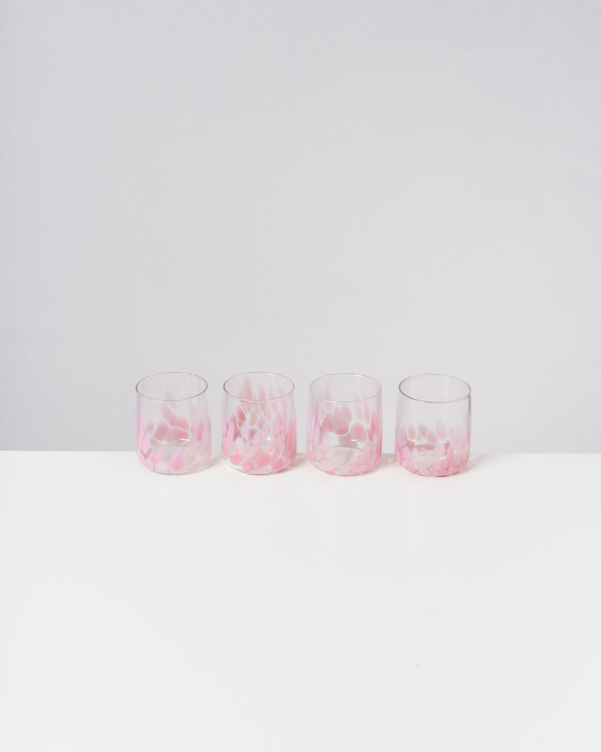 Vier rosa verzierte Gläser stehen in einer Reihe auf einer weißen Oberfläche vor einem hellgrauen Hintergrund.