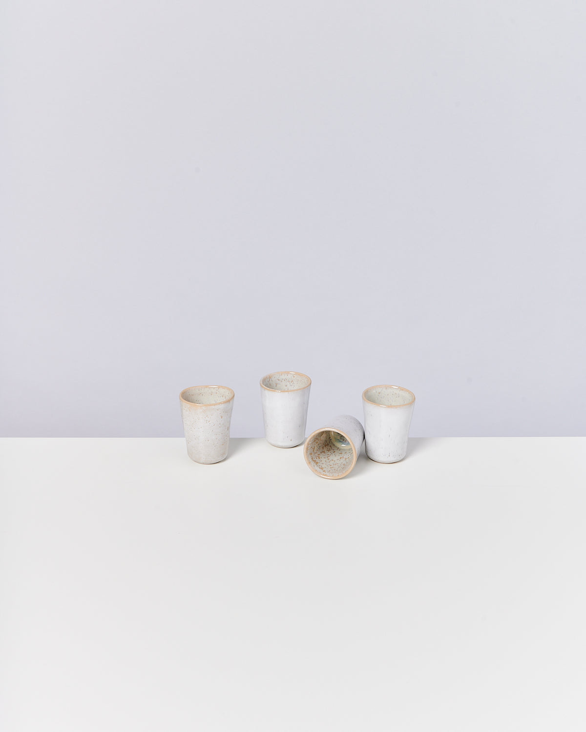 Vier weiße, handgefertigte Keramikbecher stehen und liegen auf einem weißen Tisch mit grauem Hintergrund.