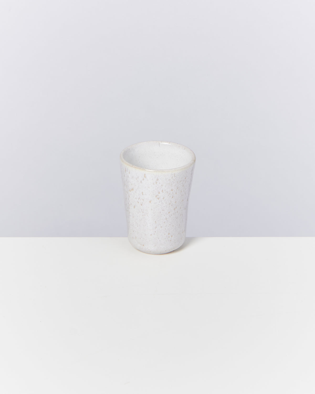 Weißer Keramikbecher mit gesprenkeltem Muster steht auf einem weißen Tisch vor grauem Hintergrund.