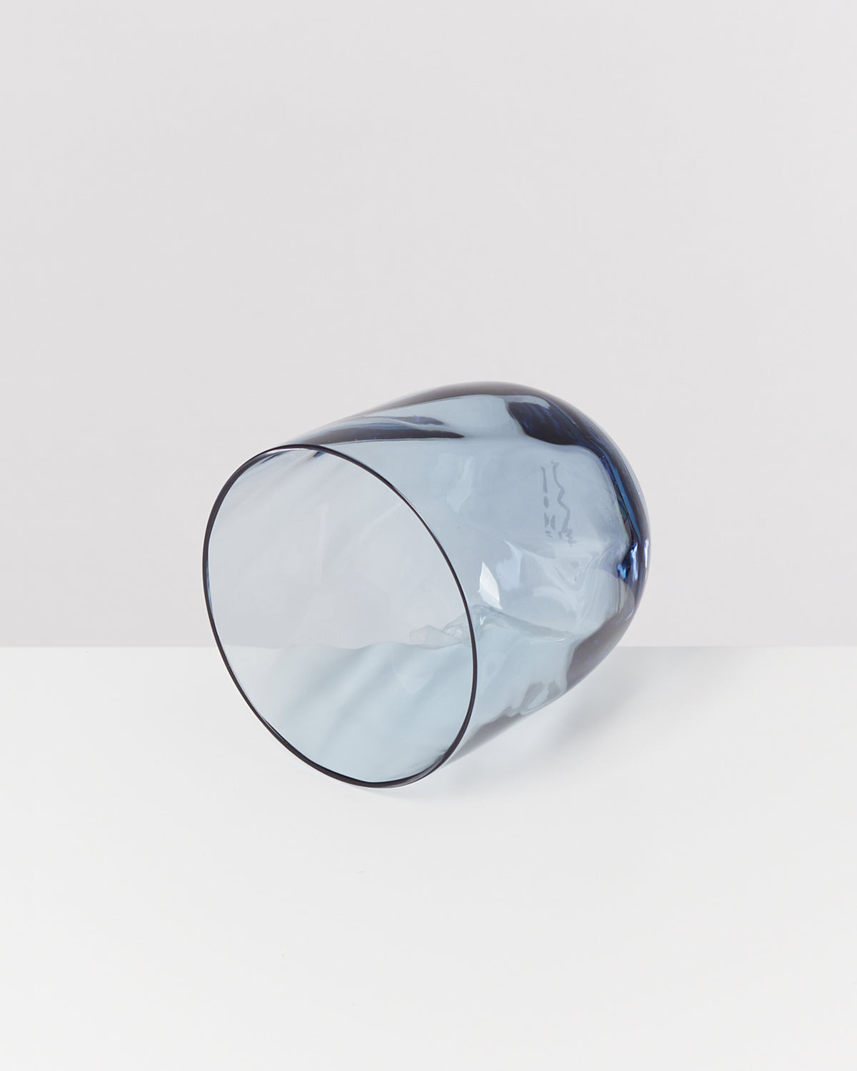 Ein transparenter, blauer Glasbecher liegt seitlich auf einer weißen Oberfläche.