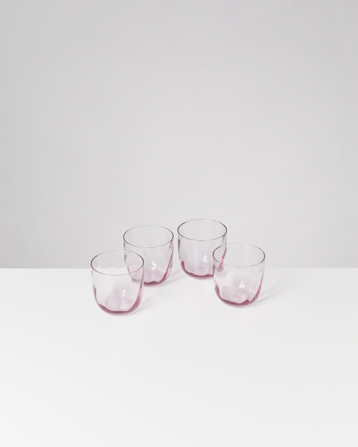 Vier durchsichtige, leicht rosa Gläser stehen auf einer weißen Oberfläche vor einem grauen Hintergrund.