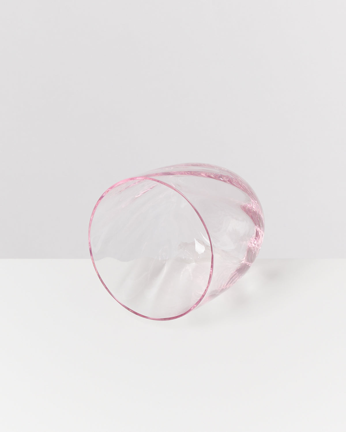 Ein rosafarbener, durchsichtiger, gewellter Glasbecher liegt seitlich auf einem weißen Hintergrund.