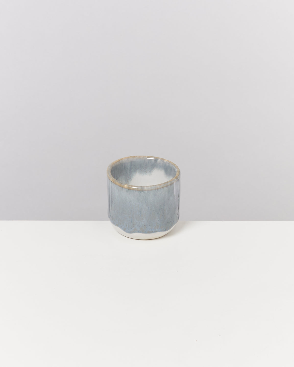 Grauer Keramikbecher mit glattem, glänzendem Finish steht auf einem weißen Tisch vor einem grauen Hintergrund.