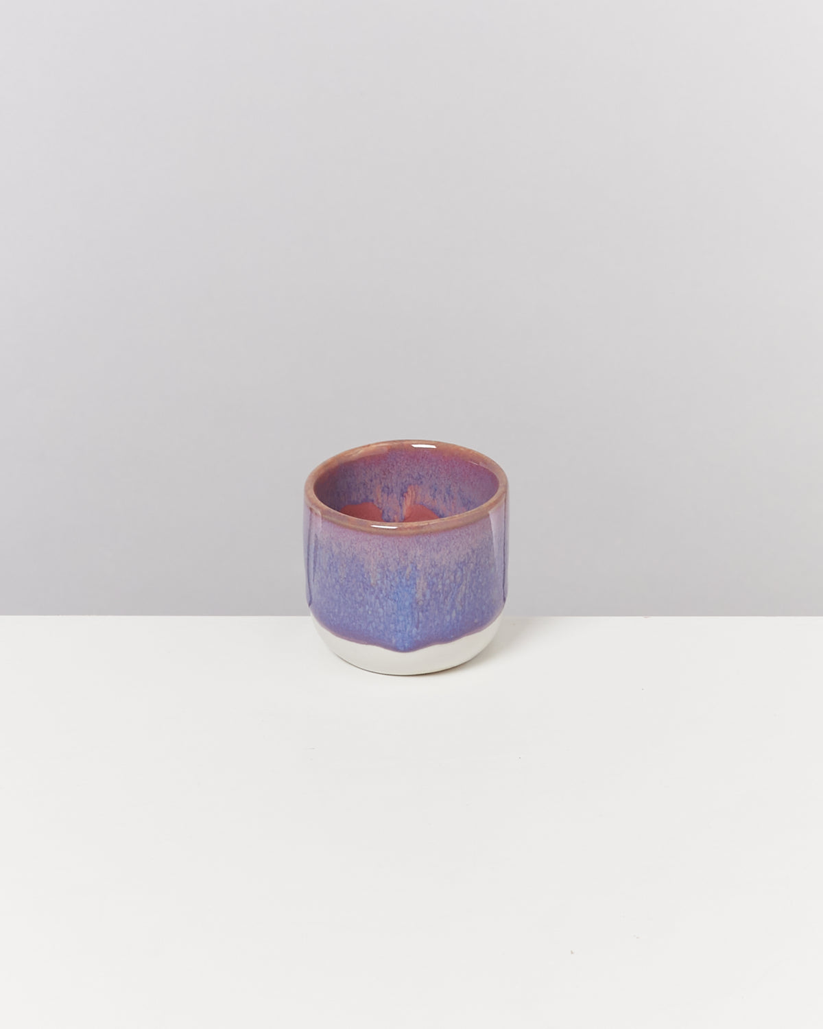 Lila-rosa Tasse mit glatter Oberfläche steht auf einem weißen Tisch vor einem grauen Hintergrund.
