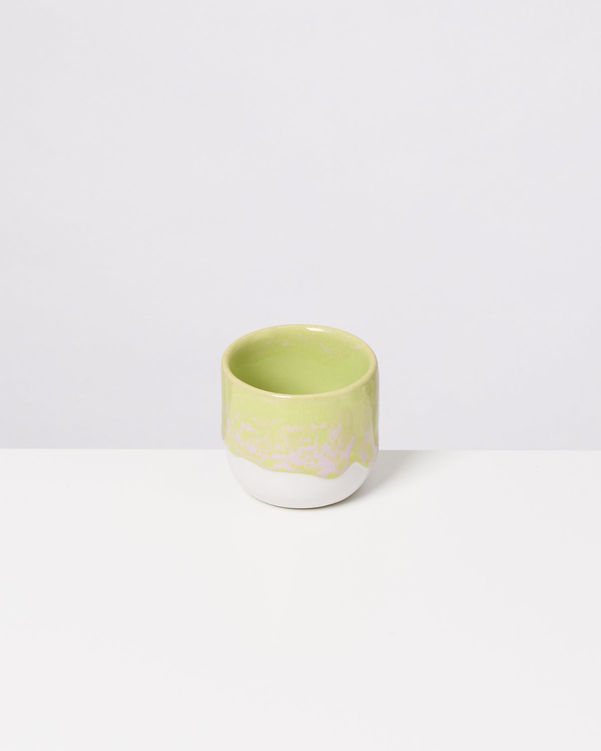 Grüne, keramische Tasse mit glatter Oberfläche steht auf einem weißen Untergrund vor einem hellen Hintergrund.