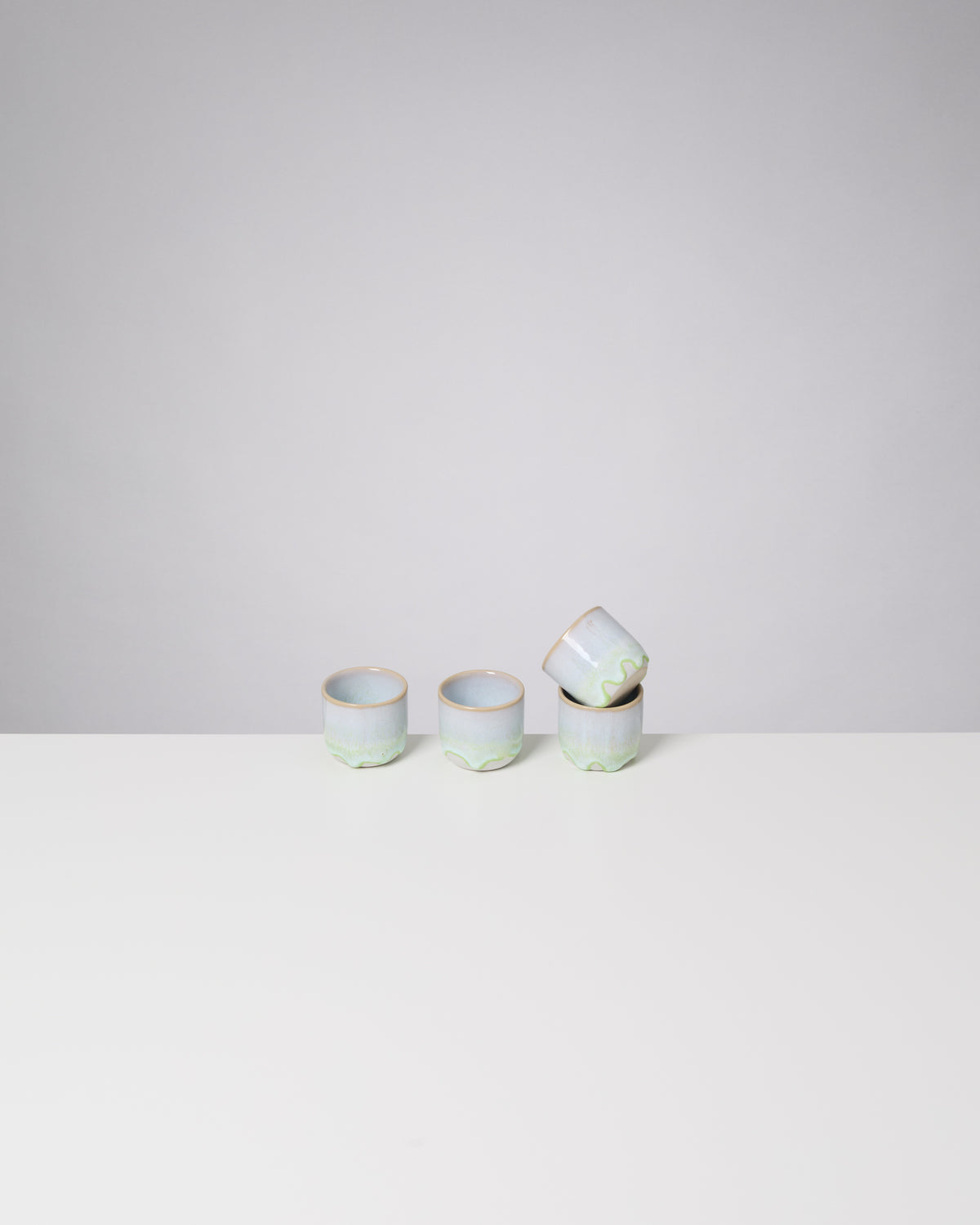 Vier kleine, pastellfarbene Tassen auf einem weißen Tisch vor grauem Hintergrund, eine Tasse liegt schräg auf der anderen.