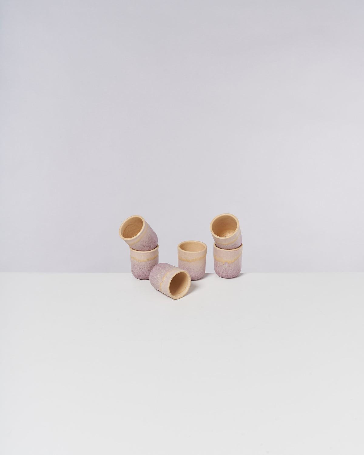 Sieben stapelbare Becher aus Ton mit einem zarten rosa Muster stehen auf einer weißen Oberfläche vor grauem Hintergrund.