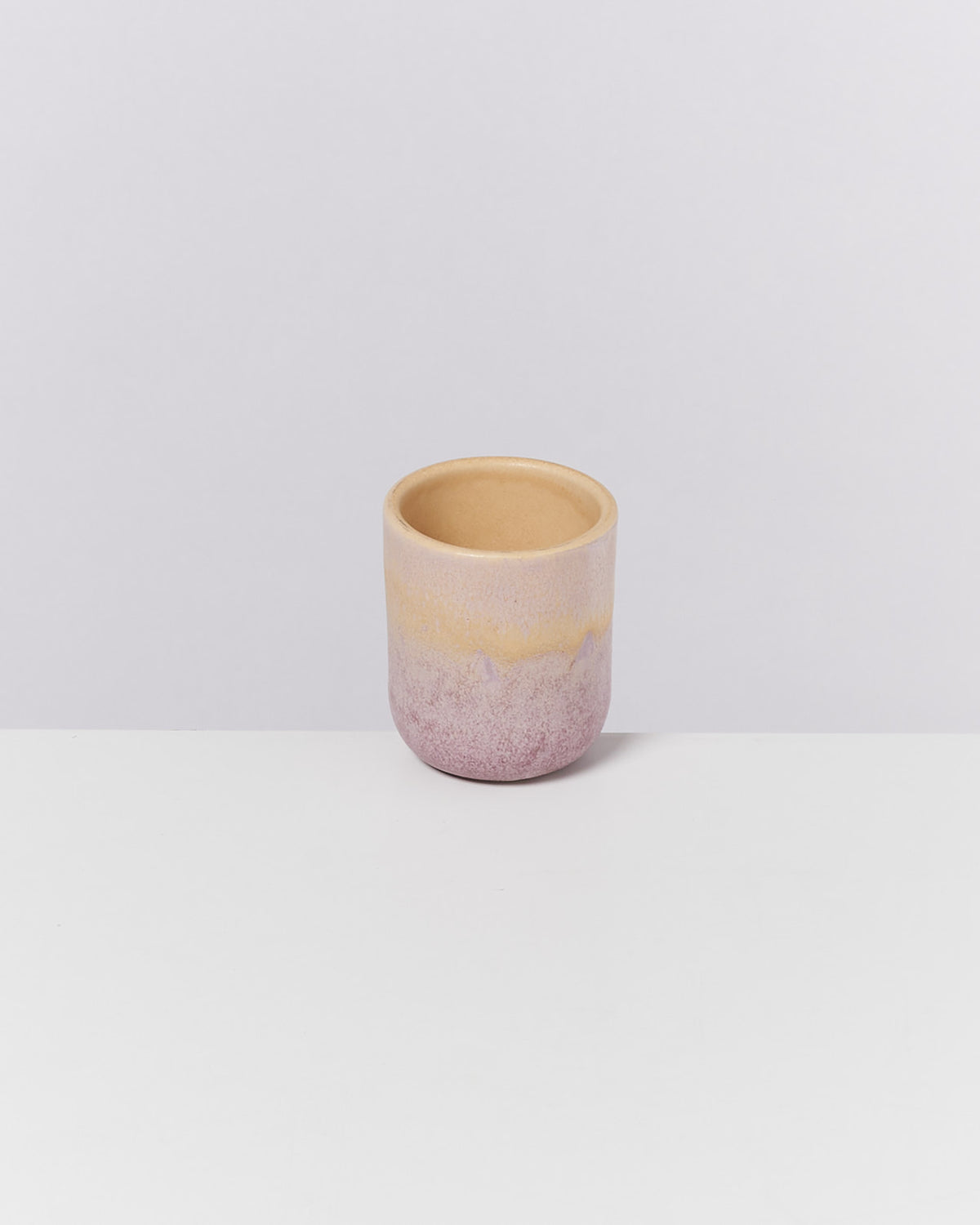 Kleiner Keramikbecher mit einem Farbverlauf von lila zu beige, auf einer weißen Oberfläche platziert.