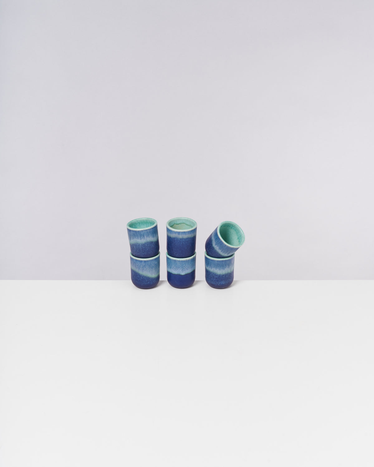 Sechs stapelbare blaue und grüne Keramikbecher auf weißem Hintergrund.