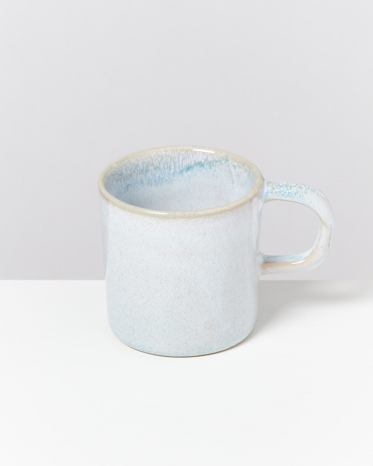 Eine hellblaue Keramiktasse mit Henkel steht auf einem weißen Tisch vor einem hellgrauen Hintergrund.