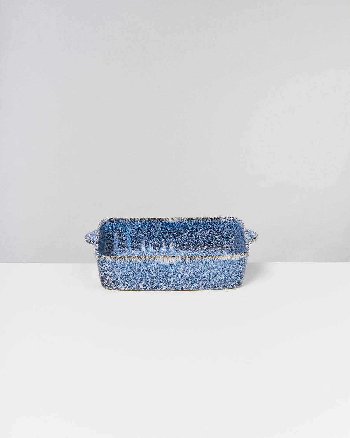 Blau gesprenkelte Keramik-Auflaufform mit Griffen auf einem weißen Tisch vor grauem Hintergrund.