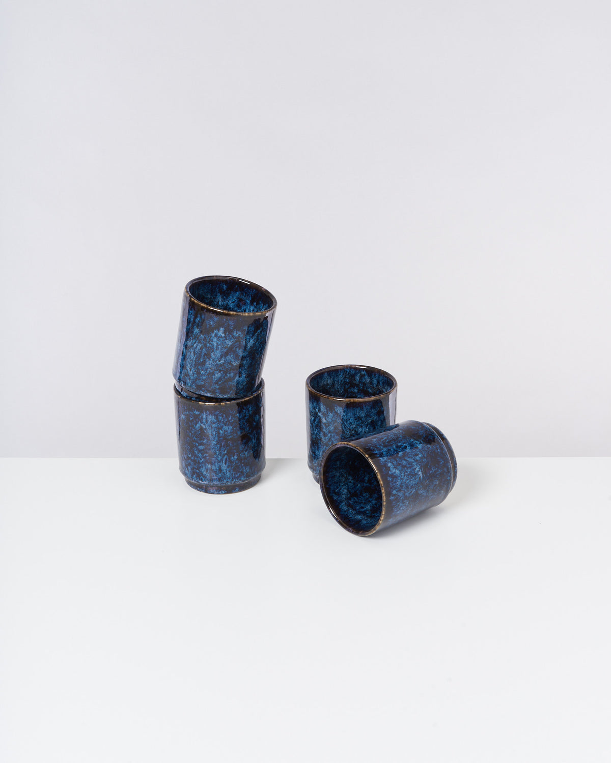Vier blaue, keramische Becher mit dunklem Muster stehen auf einem weißen Tisch.