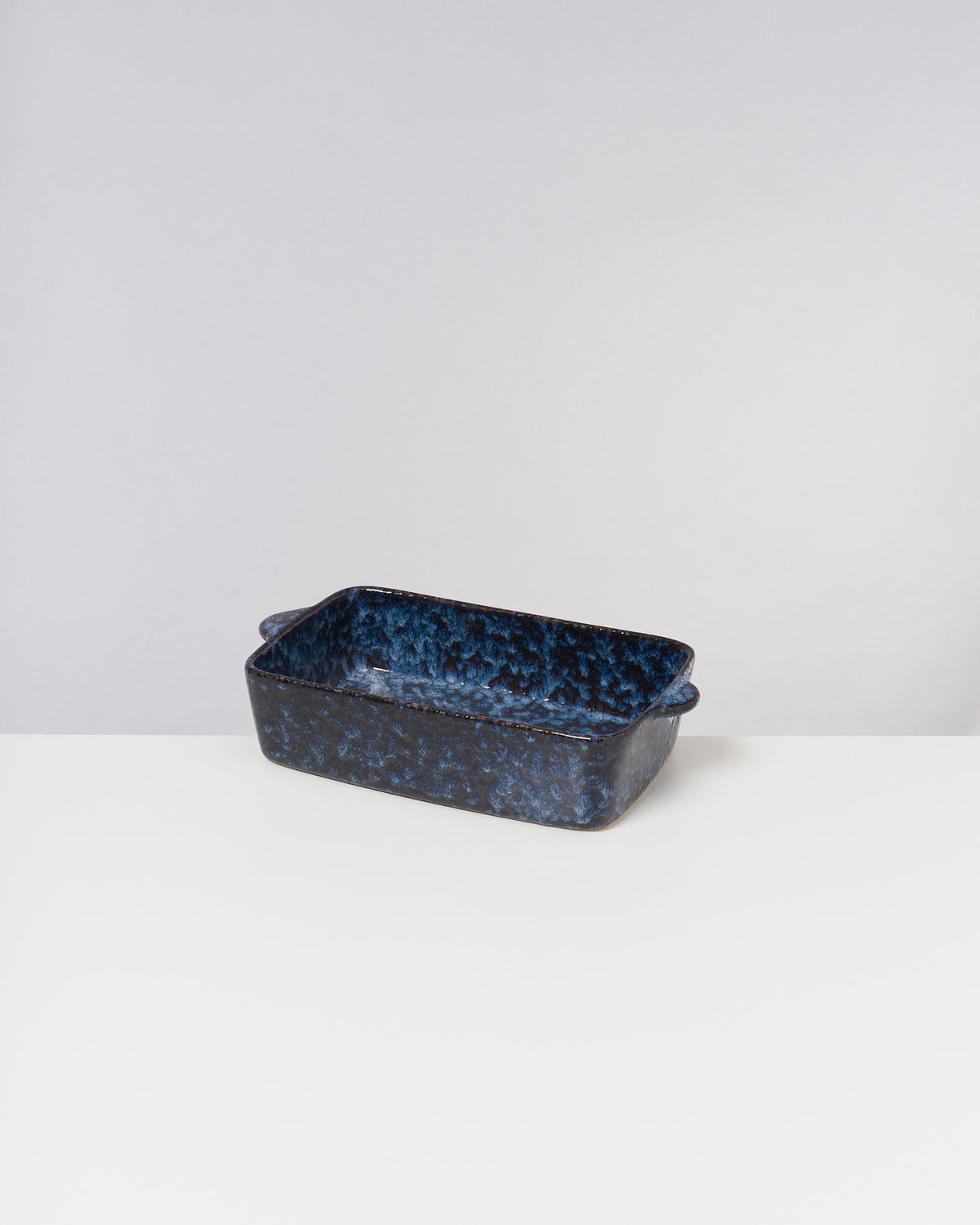 Rechteckige, blau-schwarze Keramik-Auflaufform mit Henkeln auf einem weißen Tisch vor neutralem Hintergrund.