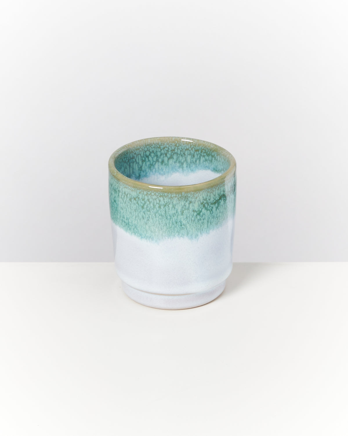 Keramikbecher mit grün-blauer Glasur auf weißem Hintergrund.