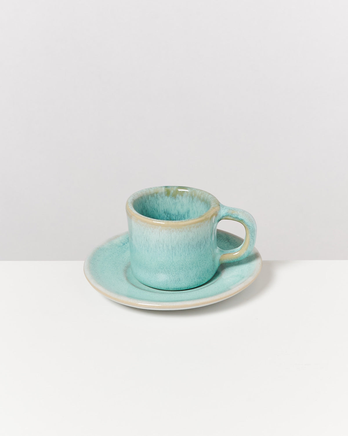 Türkise Tasse mit Untertasse auf weißem Tisch vor grauem Hintergrund.
