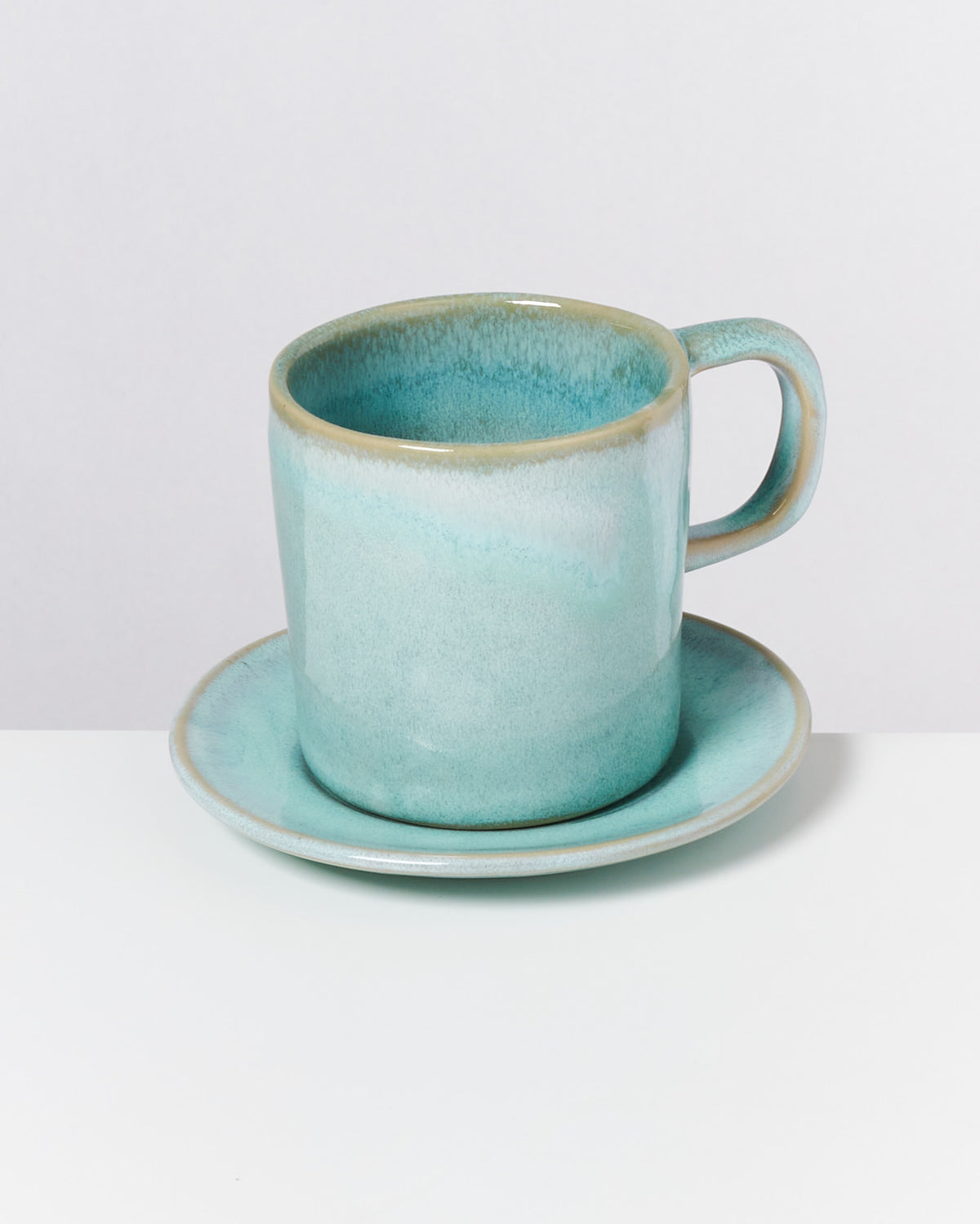 Eine türkisfarbene Keramiktasse mit Untertasse steht auf einem weißen Tisch.
