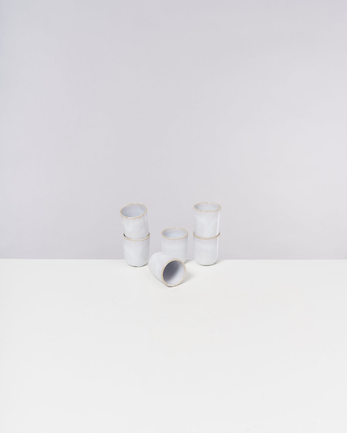 Sechs weiße, stapelbare Becher und ein umgekippter Becher auf einem weißen Tisch mit grauem Hintergrund.