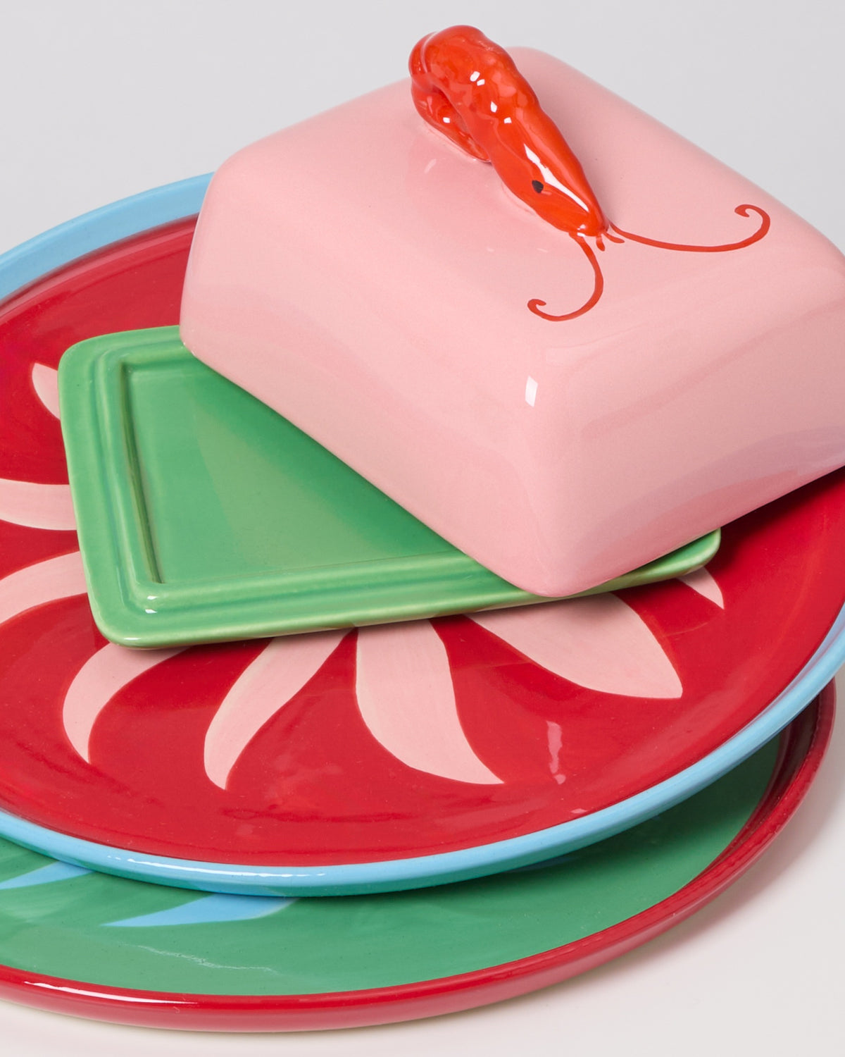 Bunte Keramikteller mit rosa Butterdose und roter Garnele als Griff, auf einem grünen Sockel platziert.
