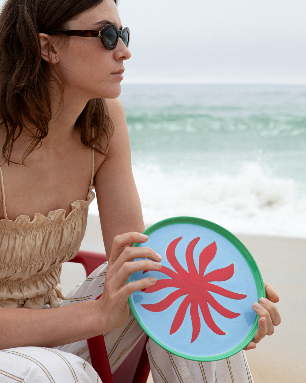 Frau mit Sonnenbrille hält einen Teller mit rotem Muster am Strand.