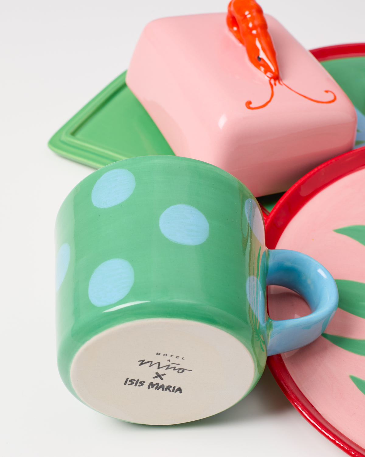 Bunte Keramikgeschirrteile: grüne Tasse mit blauen Punkten, rosa Teller mit grünem Blattmuster und rosa Butterdose.