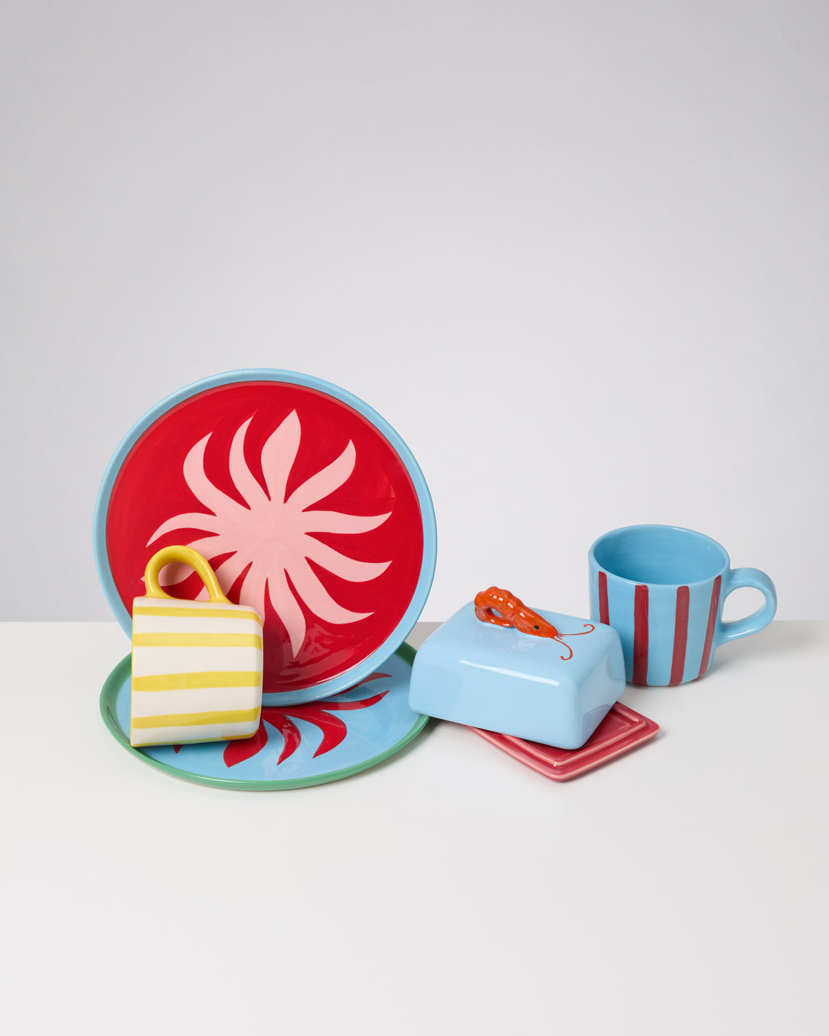 Buntes Keramikgeschirr: Teller, Tassen und Butterdose mit roten, blauen und gelben Mustern auf weißem Hintergrund.