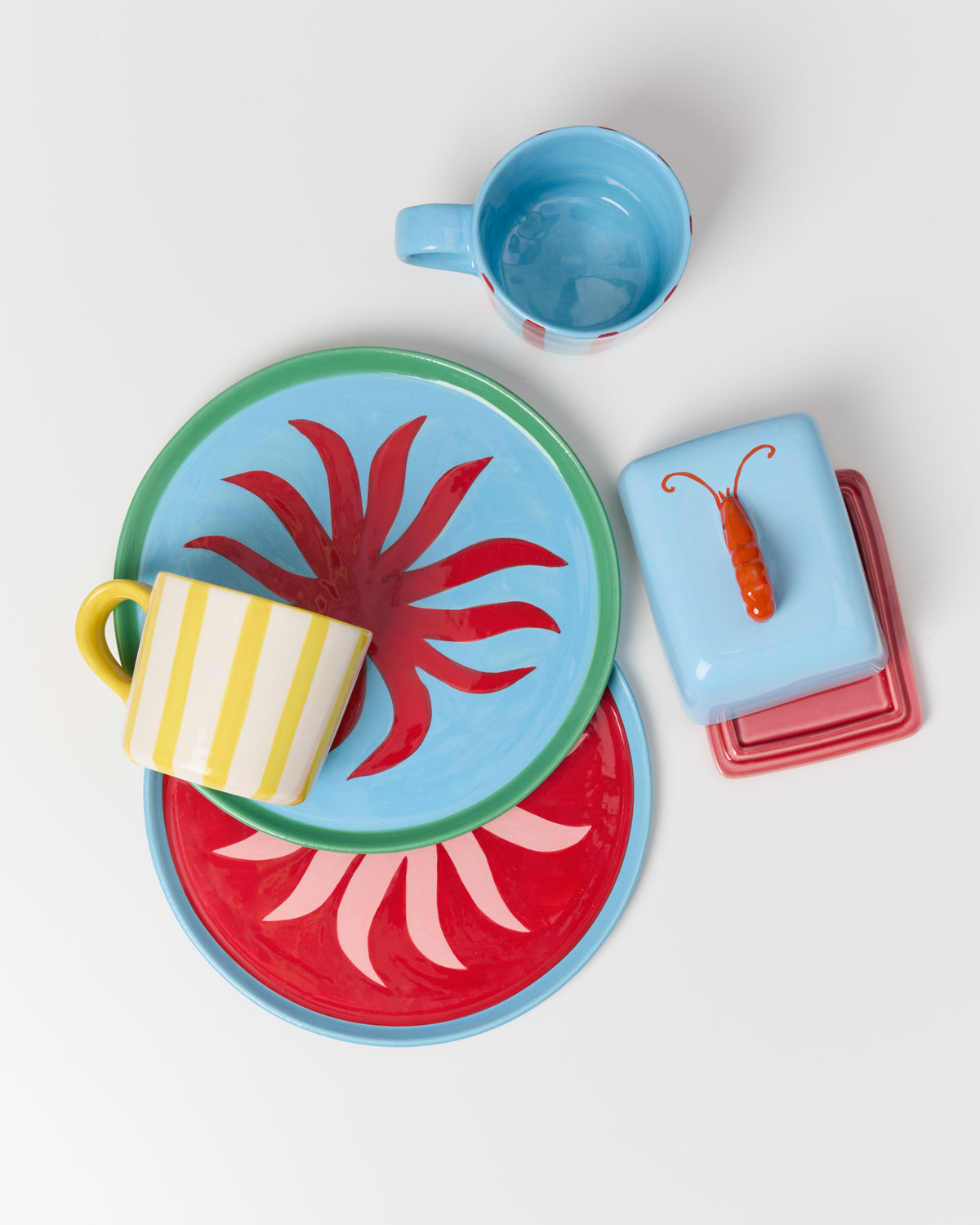Bunte Keramikteller und Tassen mit floralen Mustern und eine Butterdose mit Dekor auf weißem Hintergrund.