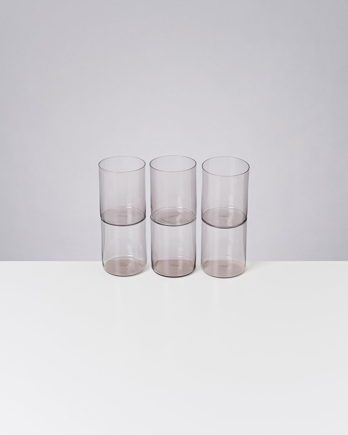 Sechs gestapelte, transparente Becher stehen auf einem weißen Tisch vor grauem Hintergrund.