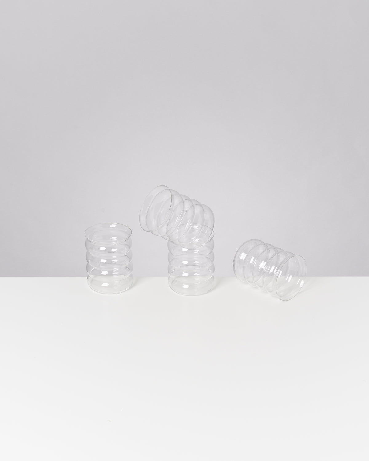 Drei durchsichtige, spiralförmige Gläser auf einem weißen Tisch, eines davon ist umgekippt.
