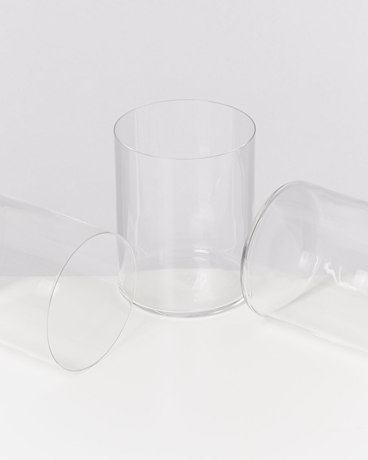 Drei leere, zylindrische Glasbecher auf einer weißen Fläche, zwei liegend, einer stehend.