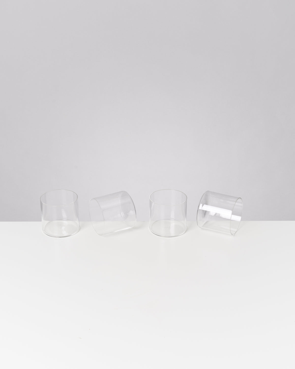 Vier transparente Gläser stehen in einer Reihe auf einem weißen Tisch, zwei davon liegen seitlich.