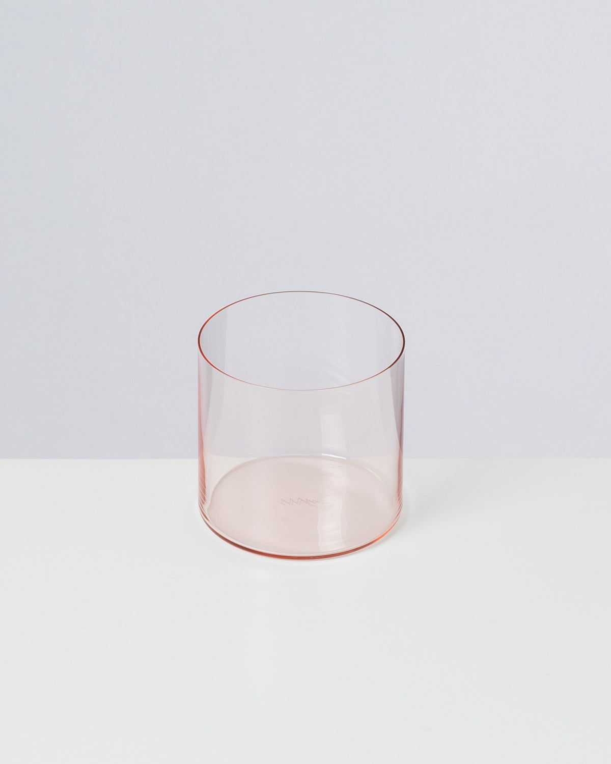 Ein durchsichtiges, leicht rosa Glas steht auf einer weißen Oberfläche vor grauem Hintergrund.