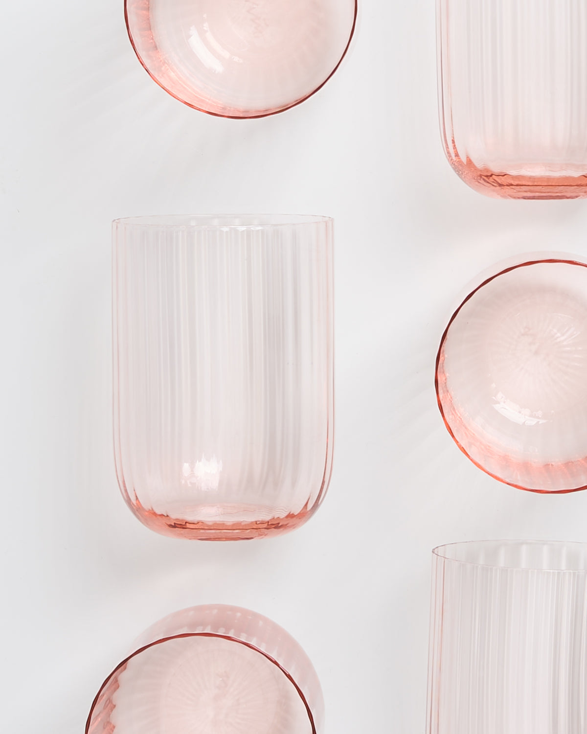 Geriffelte, roséfarbene Glasgefäße, darunter Gläser und Schalen, auf weißem Hintergrund angeordnet.