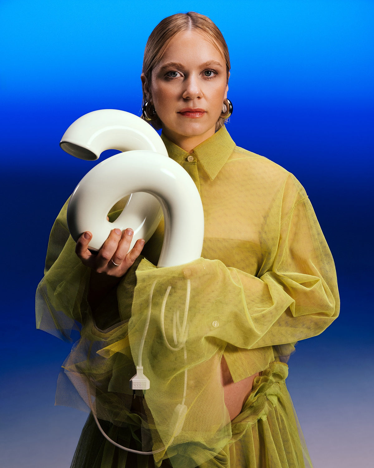 Person in gelbem Outfit hält eine weiße, skulpturale Lampe mit Kabel vor einem blauen Hintergrund.