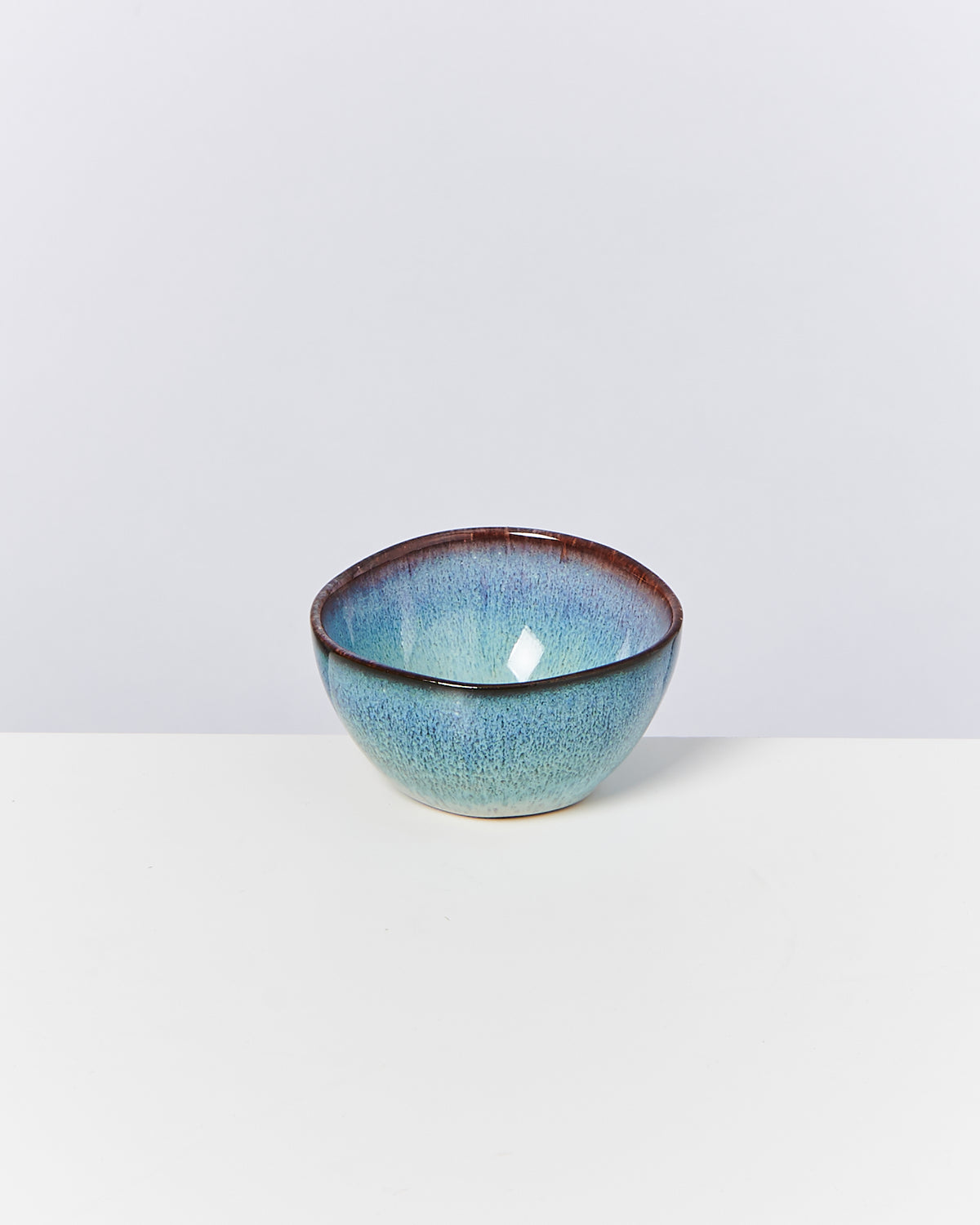 Eine kleine, blau glasierte Schüssel mit braunem Rand steht auf einem weißen Tisch vor einem grauen Hintergrund.