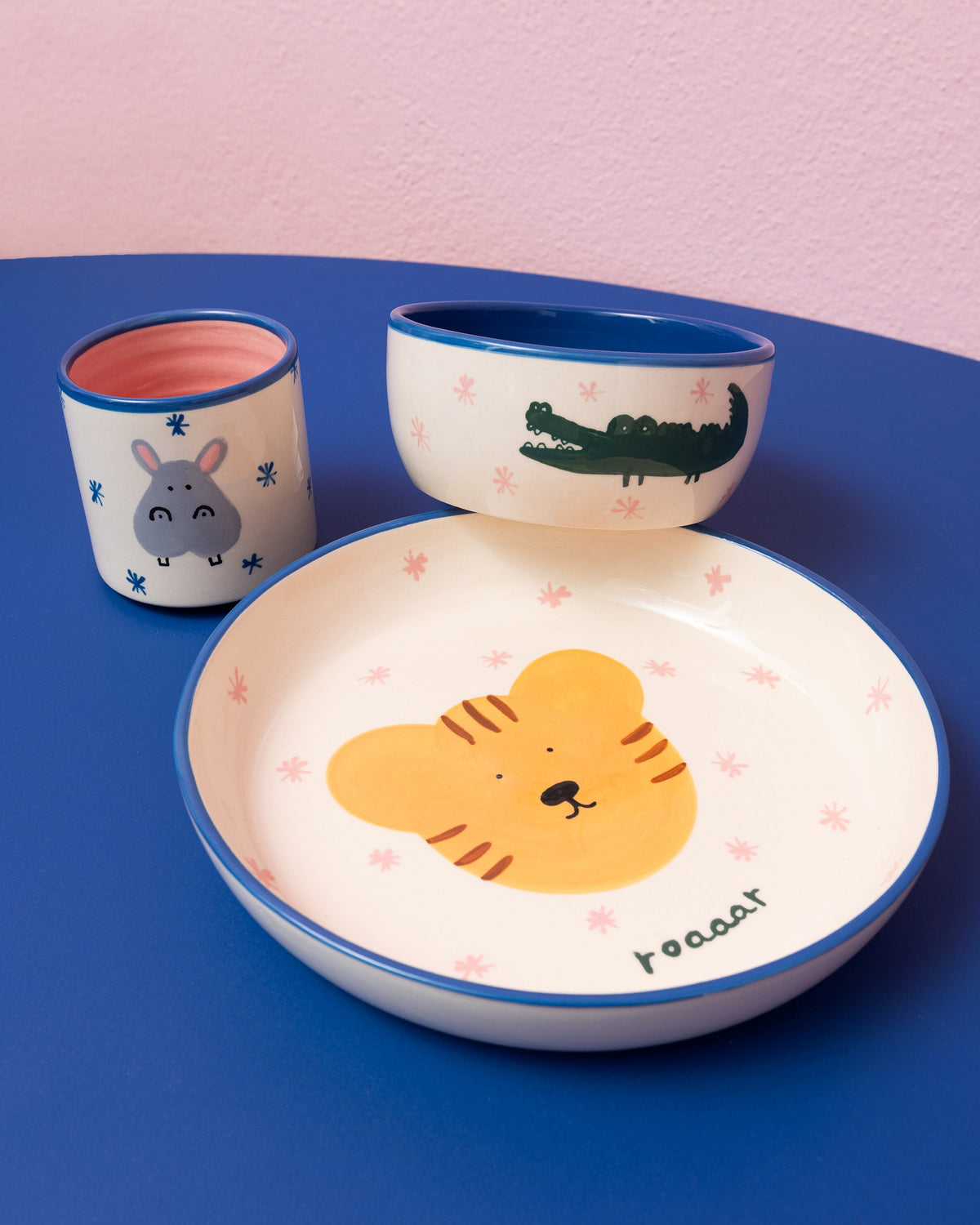 Kindergeschirr-Set mit Tiermotiven: Teller mit Tiger, Schale mit Krokodil und Becher mit Nilpferd auf blauem Tisch.