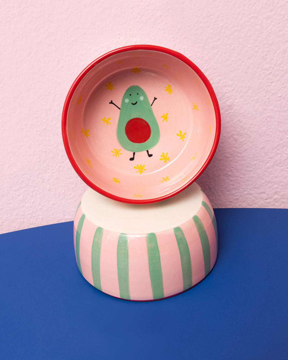 Bunte Schale mit Avocado-Motiv und Blumen auf gestreifter Basis vor rosa Hintergrund.