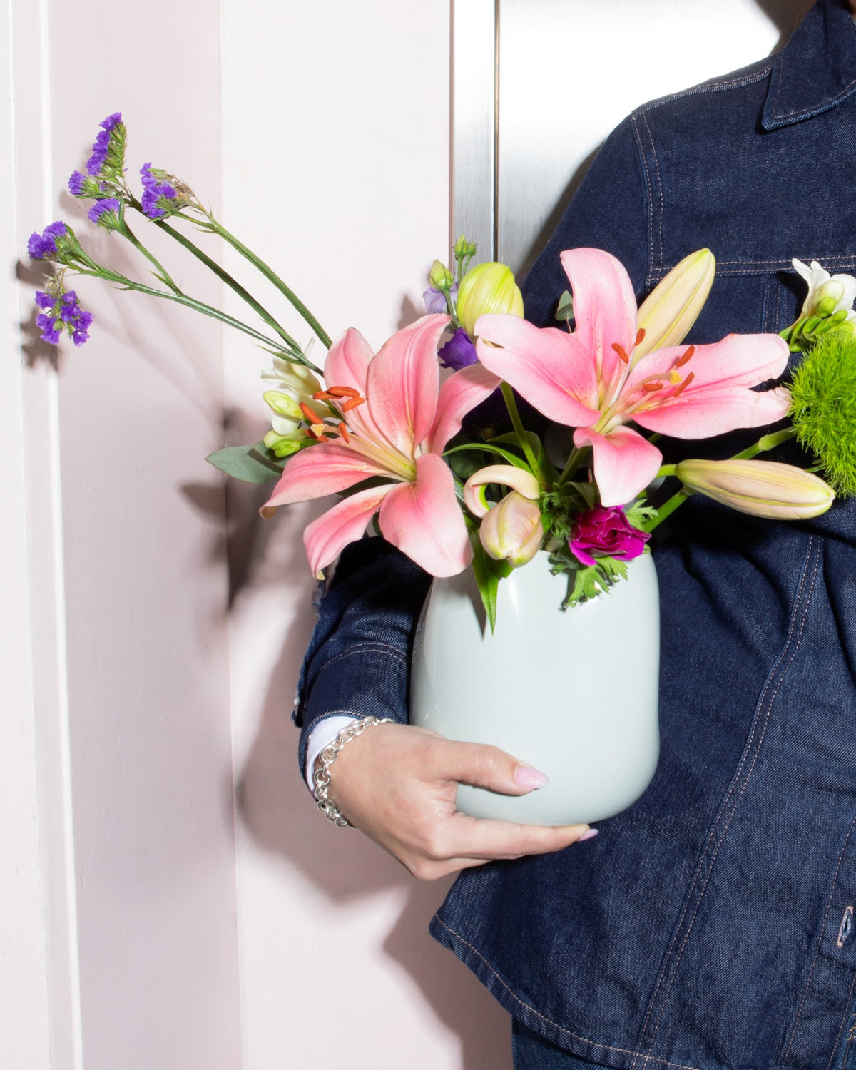 Person in Jeansjacke hält eine Vase mit rosa Blumen, darunter Lilien, und violetten Blüten vor pastellfarbenem Hintergrund.