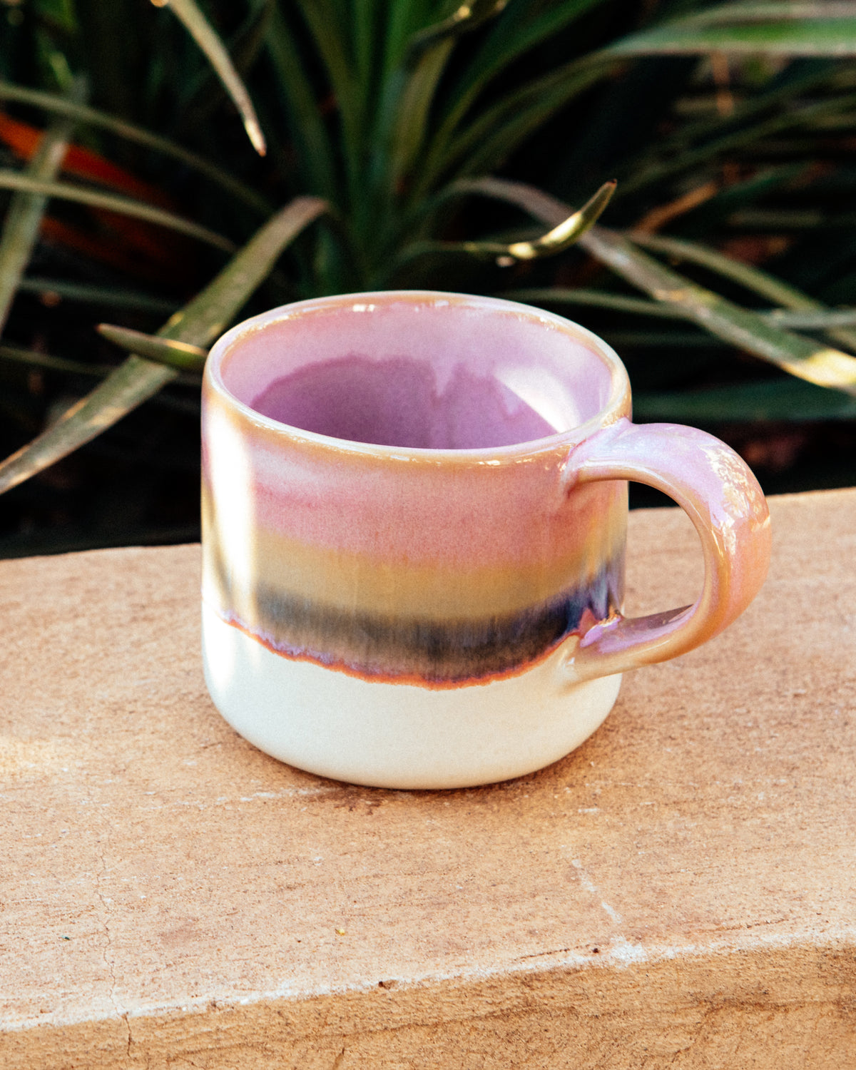 Keramiktasse mit rosa, gelben und violetten Streifen steht auf einem Ziegelstein, im Hintergrund Pflanzen.
