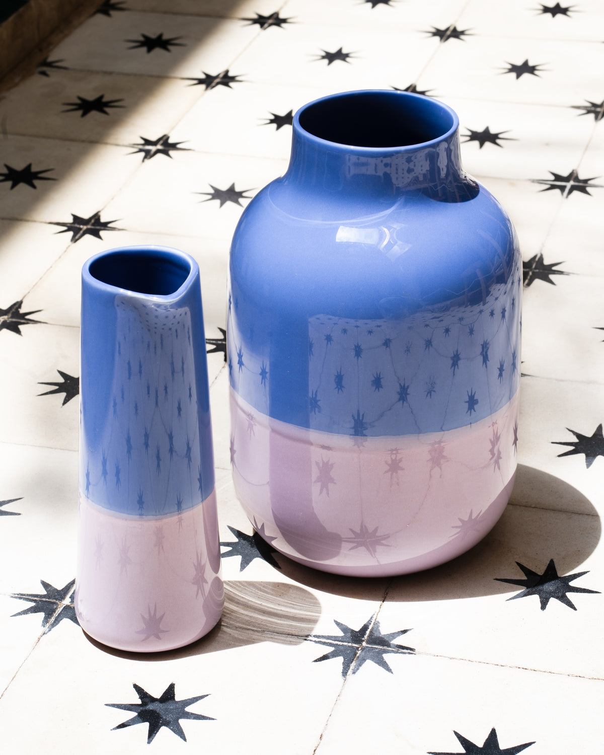 Zwei Keramiken in Blau und Rosa stehen auf einem Boden mit sternförmigen Mustern im Sonnenlicht.