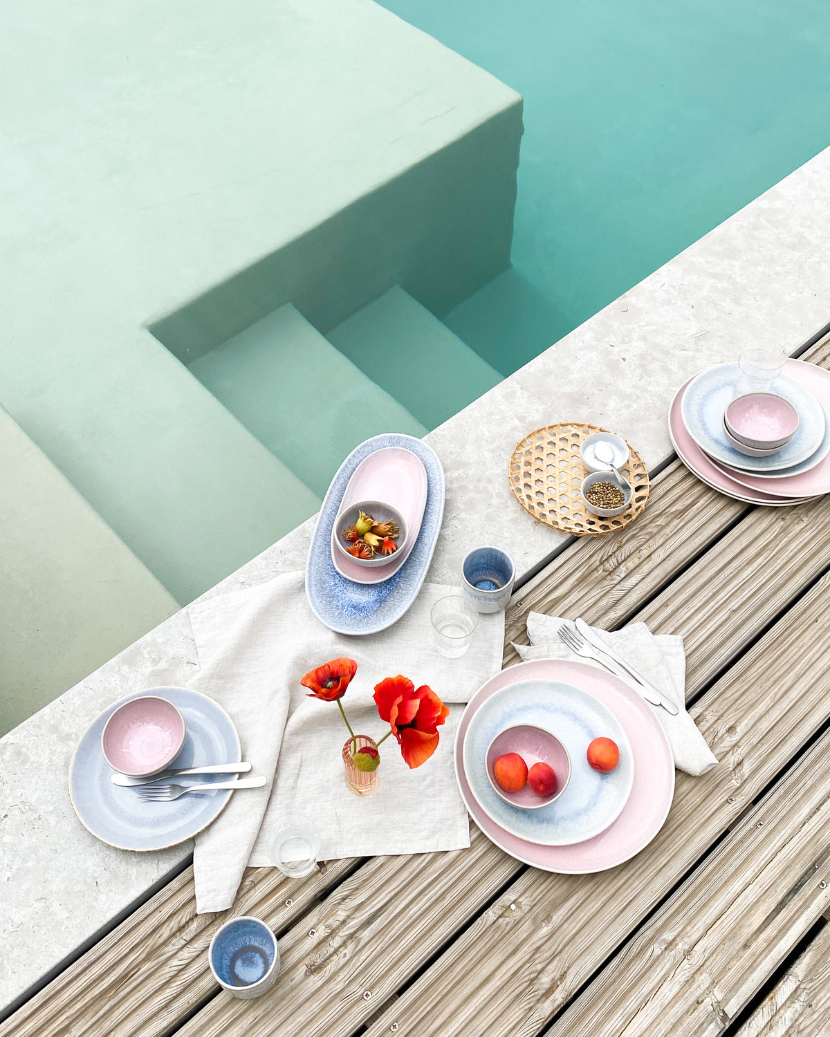 Ein gedeckter Tisch mit buntem Geschirr und Blumen am Rand eines Pools.