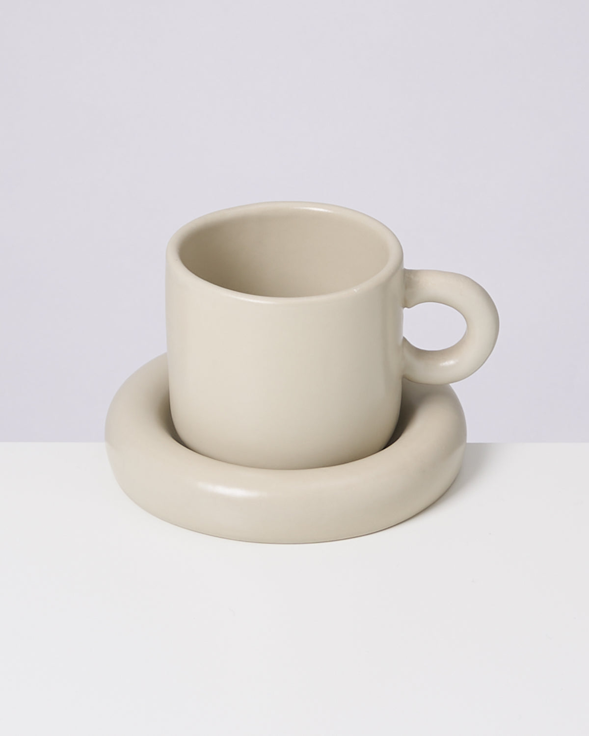 Beige Tasse mit rundem Henkel steht in passendem, gerundetem Untersetzer auf weißem Untergrund.
