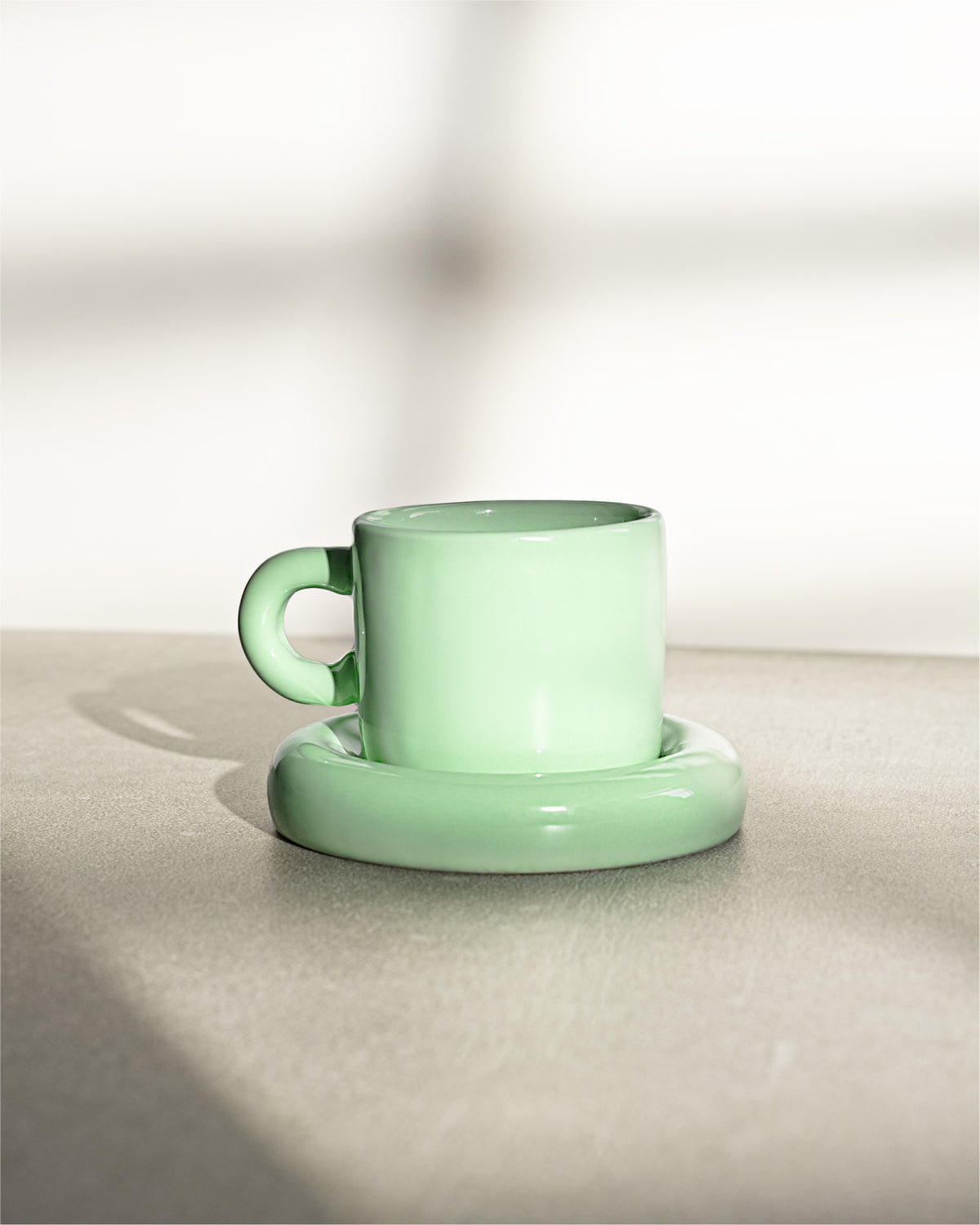 Grüne Tasse mit Untertasse auf einem neutralen Untergrund, im Sonnenlicht fotografiert.
